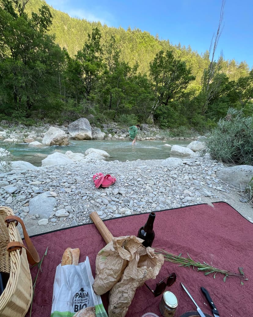 A midsummer picnic on a burgundy blanket by a mountain river — baguette, wild herbs, glass bottle and wicker basket in the foreground, a figure wading across the shallow stream beyond.