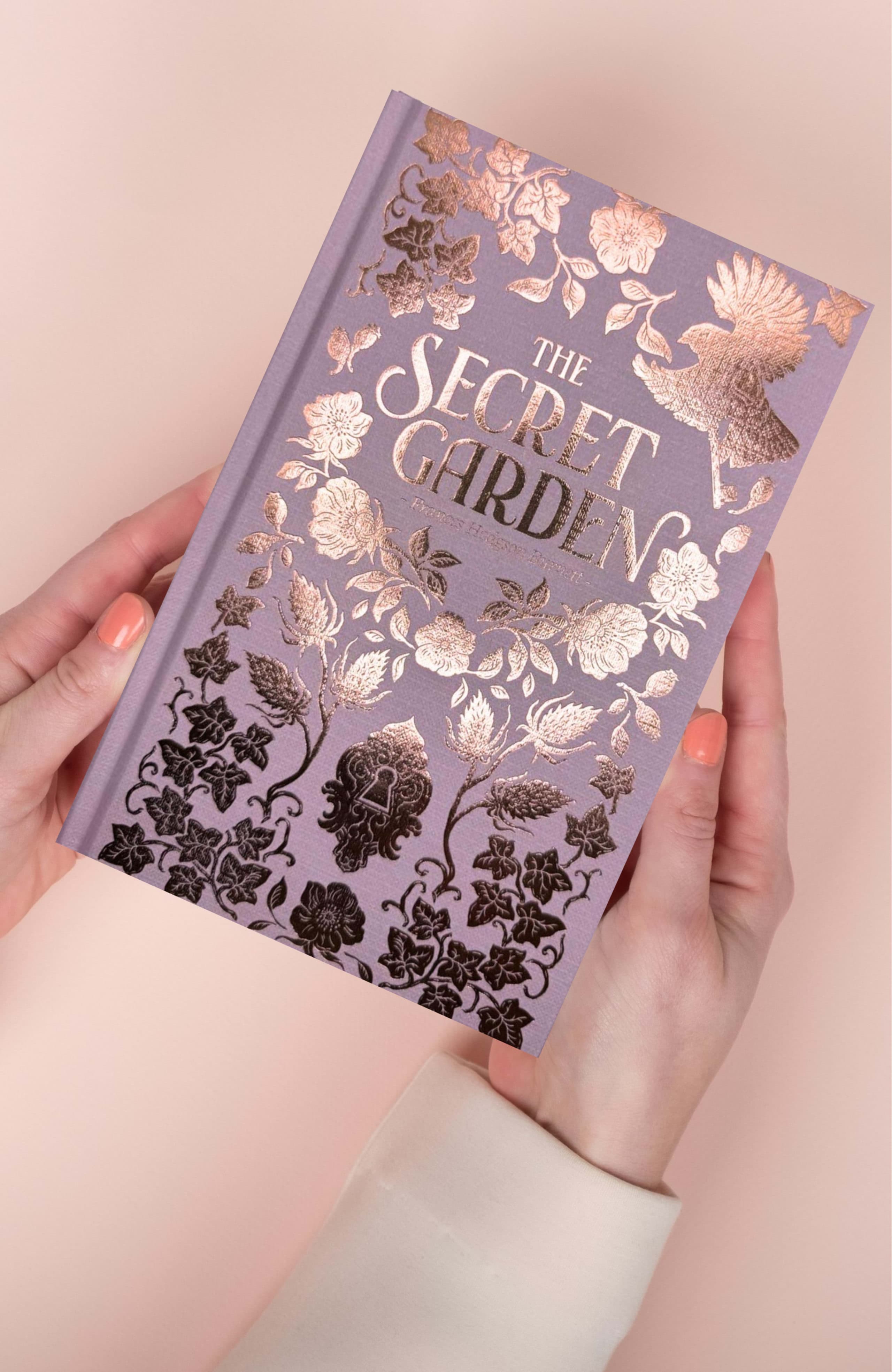 Hardback book cover in lavender cloth held in two hands, with copper and black foil-stamped botanical ornament and title text.